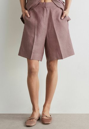 Kvinde iført mauve knælange skræddersyede shorts med lommer og matchende balletsko, stående mod en ensfarvet baggrund.