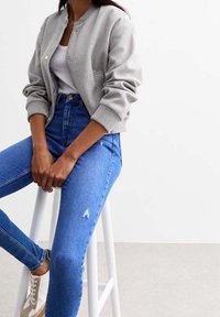 Mujer sentada en un taburete blanco que lleva una chaqueta bomber gris clara, una camiseta blanca sin mangas, vaqueros de tiro alto azules y zapatillas beige.