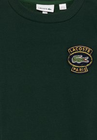 Sudadera verde oscuro hecha de una tela suave. Presenta un parche bordado del logo de Lacoste con un cocodrilo y "PARÍS" en el pecho.