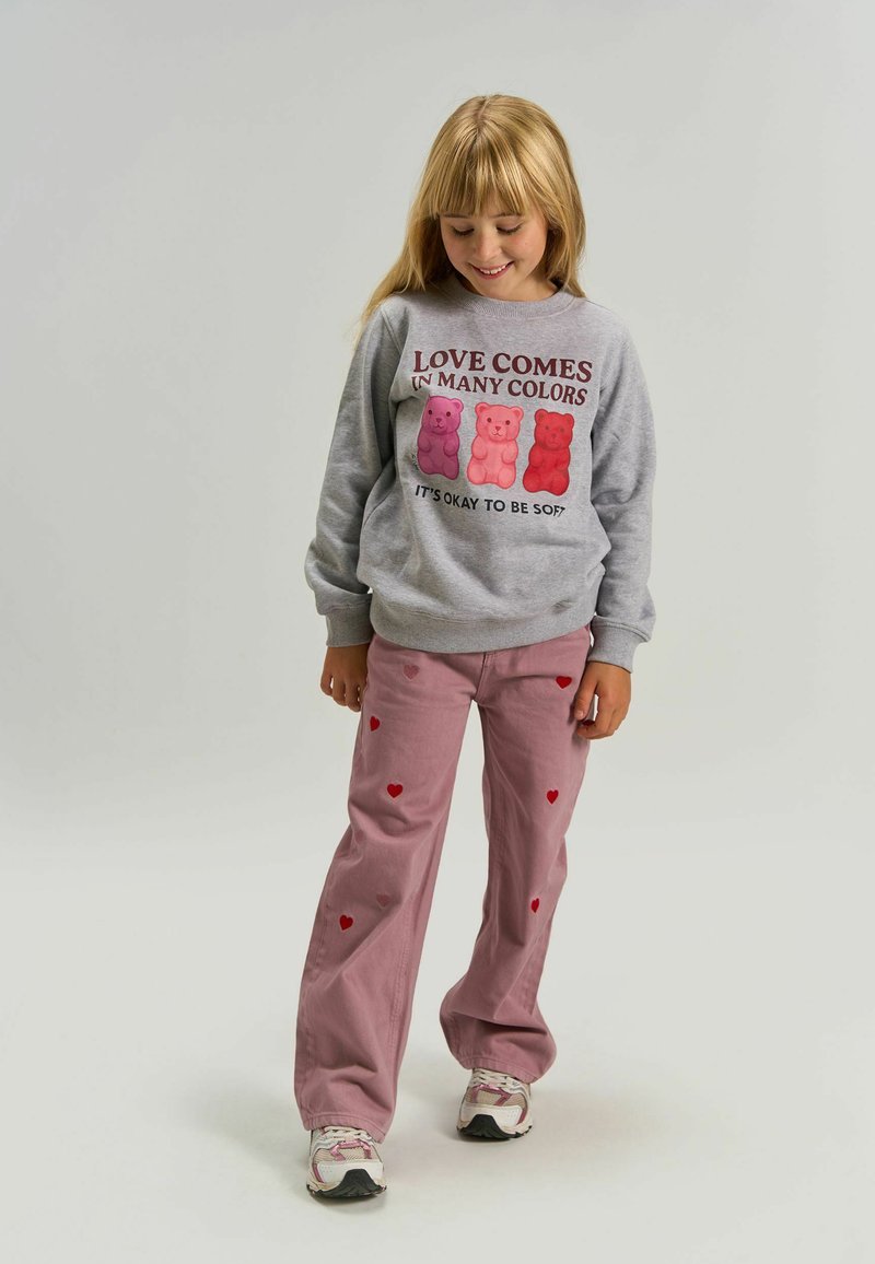 Jeune fille portant un sweat-shirt gris avec des ours roses et du texte, un pantalon rose avec des cœurs rouges, et des baskets blanches, debout et souriante.
