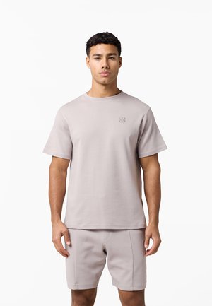 Giovane uomo in piedi che indossa una t-shirt beige chiaro a maniche corte e pantaloncini abbinati, rivolto verso l’obiettivo su uno sfondo bianco neutro.