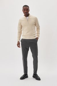 Beige, cipzáros pulóver, puha textúrával, szürke, szabott nadrággal és fekete, slip-on cipővel párosítva. A modell sima háttér előtt áll.