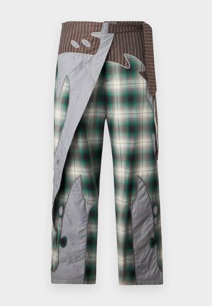 Joven con chaqueta negra acolchada, camisa oscura, pantalones verdes de cuadros grandes con detalles en tela gris y zapatillas blancas, de pie.