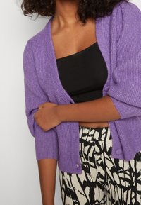 Cárdigan de punto morado con mangas cortas, con un cierre de botones y tejido texturizado; combinado con pantalones de estampado en blanco y negro.
