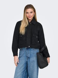 Jeune femme aux longs cheveux blonds portant une veste noire, un jean large bleu et tenant un sac à main noir sur le bras, sur un fond clair.