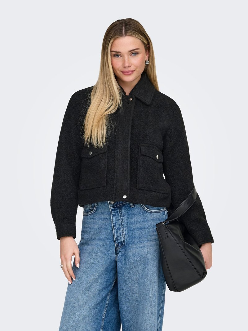 Jeune femme aux longs cheveux blonds portant une veste noire, un jean large bleu et tenant un sac à main noir sur le bras, sur un fond clair.