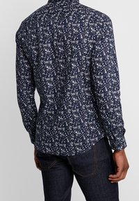 Camisa de manga larga en tejido azul marino oscuro con un patrón de paisley blanco. Cuenta con un cuello abotonado y un dobladillo curvado. Textura suave, diseño ajustado.