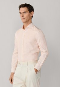 Mężczyzna w jasnoróżowej koszuli w paski z długim rękawem, wpuszczonej w beżowe spodnie z wysokim stanem, stojący z jedną ręką w kieszeni.