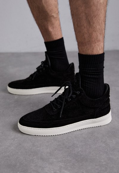 Sorte semskede sneakers med teksturert overflate, med hvit gummisåle, sorte lisser og polstret krage, mot en grå bakgrunn.
