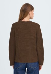 Brauner strukturierter Strickpullover mit Rundhalsausschnitt und geripptem Saum, der eine lockere Passform und fallende Schultern aufweist. Sichtbares Strickmuster auf dem Stoff.
