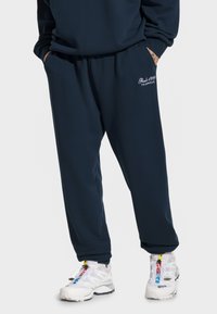 Granatowe dresy wykonane z miękkiego materiału, z luźnym krojem i elastycznymi mankietami. Napis "Bashi 1999 Teamwear" w białym kolorze na lewej uldze.