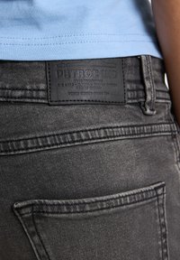 Jeans de mezclilla negros con una etiqueta de cuero visible que lleva la marca "PETROL IND" y un diseño de un solo bolsillo trasero. Textura suave y acabado desvaído.