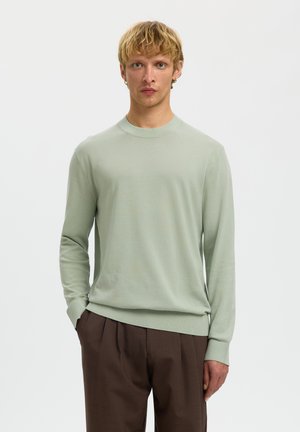 Hombre rubio que lleva un jersey de manga larga verde claro y pantalones plisados marrón oscuro, de pie con una mano en el bolsillo contra un fondo liso.