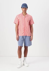 Stribet kortærmet skjorte i koral- og hvid, parret med lyseblå shorts. Hvide sneakers og en blå kasket fuldender det afslappede look.