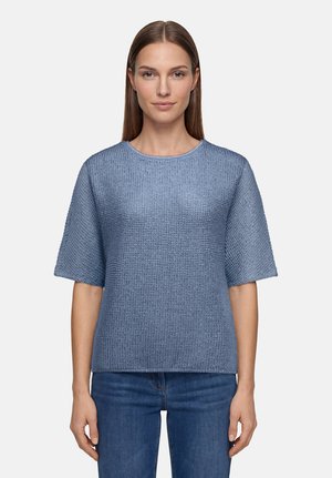 Femme aux cheveux bruns et raides porte un pull bleu texturé à manches courtes et un jean bleu, debout devant un fond clair uni.