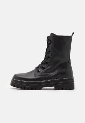 Botte de combat noire en cuir à lacets avec semelle épaisse, tige montant à la cheville, œillets métalliques et languette arrière, sur un fond clair uni.