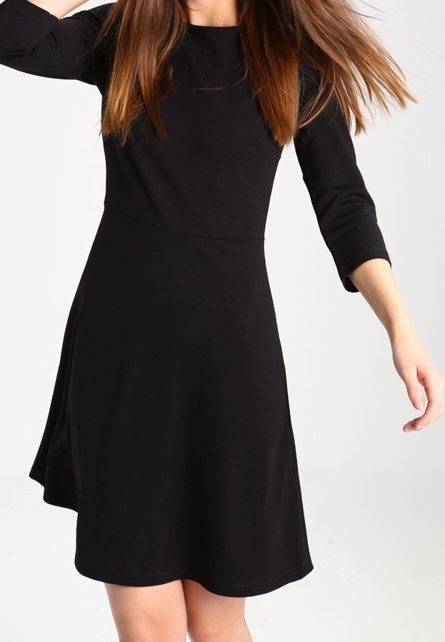 Femme portant une robe noire ajustée avec des manches trois-quarts, debout avec un bras levé et l'autre tendu vers l'extérieur.