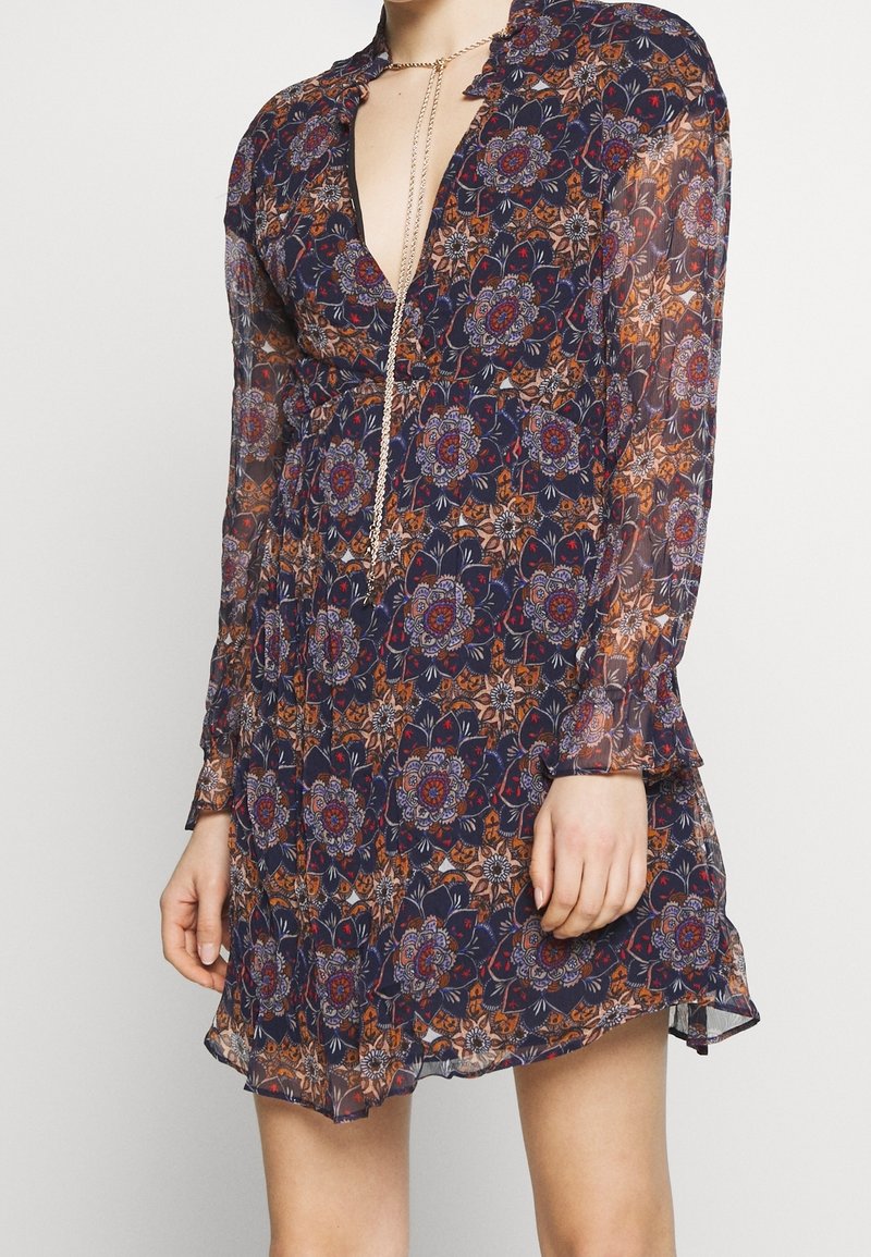 Robe florale bleu marine avec manches longues en tissu transparent, décolleté en V profond, col à volants et ourlet fluide. Présente des motifs multicolores complexes.