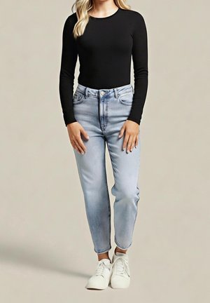 Kvinde iført en sort langærmet top, lyseblå højhælede jeans og hvide sneakers, der står mod en ensfarvet baggrund.