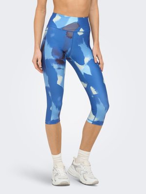 Frau trägt hoch taillierte blaue gemusterte kurze Leggings, weiße Socken und weiße Sportschuhe, steht vor einem einfarbigen Hintergrund.