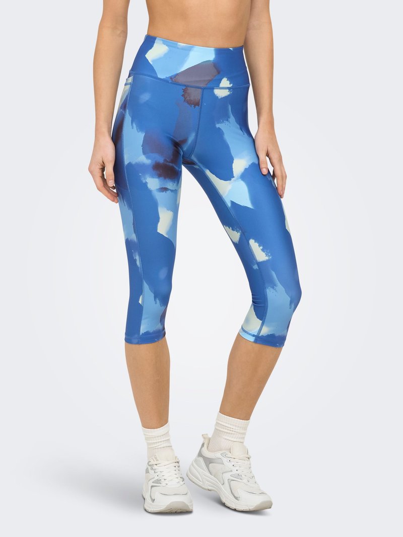 Femme portant des leggings courts bleu à motifs taille haute, des chaussettes blanches et des chaussures de sport blanches, debout devant un fond uni.