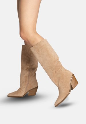 Piernas con botas de ante color marrón claro hasta la rodilla, con cremalleras laterales y tacones gruesos apilados de altura media, sobre un fondo claro.