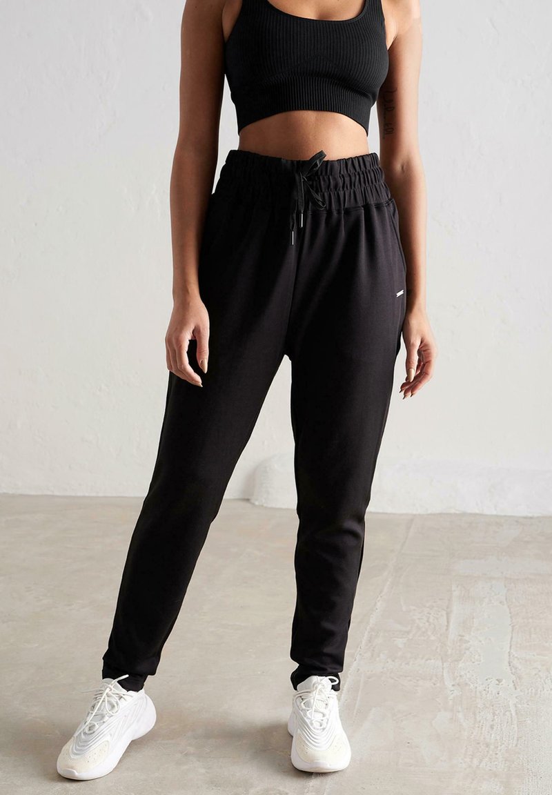 Sorte joggers lavet af blødt stof med elastisk talje, justerbar snor og sidelommer, parret med hvide sneakers.