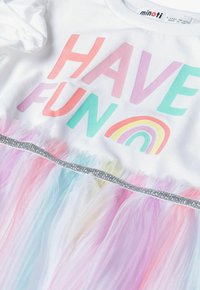 T-shirt blanc avec le texte coloré "AMUSEZ-VOUS", mettant en avant un graphique arc-en-ciel. Jupe en tulle pastel superposé avec une ceinture en paillettes argentées.