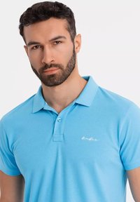 Ljusblå pikétröja med texturerad tyg, korta ärmar, krage och två knappar. Har en liten vit logotyp på bröstet.