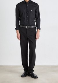Uomo in piedi con una camicia nera aderente, cintura nera, pantaloni neri su misura e mocassini in pelle nera su uno sfondo semplice.
