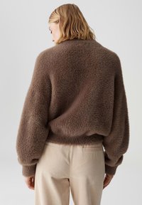Vrouw in een bruine pluizige trui met ballonmouwen en crèmekleurige corduroy broek, staande met haar rug naar de camera tegen een effen achtergrond.