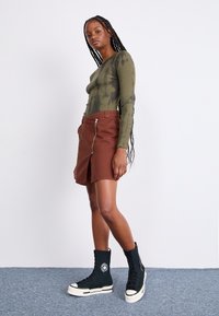 Haut à manches longues tie-dye vert, jupe devant zippée couleur rouille, et baskets montantes noires avec semelles blanches et patch logo, posant devant un fond clair.