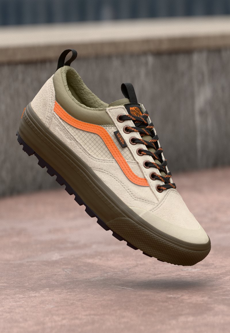 Zapatilla beige con lona texturizada y acentos de goma lisa, con una franja lateral naranja, cordones negros y una suela morada robusta.