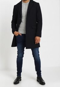 Cappotto di lana blu navy con colletto a punte, bottoni frontali, tasche laterali e un maglione grigio a trecce sotto, abbinato a jeans scuri e scarpe nere.