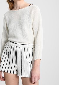 Vit stickad tröja med vid halsringning, kombinerad med vita shorts med svarta vertikala ränder och elastisk midja.