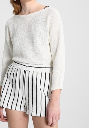 Maglione bianco lavorato a maglia con scollo ampio, abbinato a pantaloncini bianchi con strisce verticali nere e un elastico in vita.