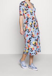Blumenkleid aus hellblauem Stoff mit gelben und roten Obstmotiven, kurzen Ärmeln und tiefem V-Ausschnitt. Kombiniert mit hellblauen Turnschuhen.