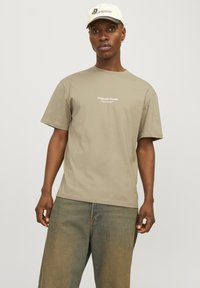 Khakifarbenes T-Shirt mit kurzen Ärmeln und Rundhalsausschnitt, auf der Brust mit "Originals Studio" in Weiß bedruckt, kombiniert mit faded weit geschnittenen Jeans.