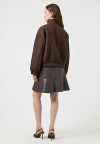 Chaqueta bomber marrón acolchada con textura floral, puños y dobladillo elásticos, llevada sobre una falda de cuero oscura. Tacones abiertos en la punta complementan el conjunto.