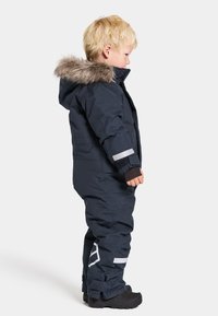 Mörkblå barnoverall med huva fodrad med syntetisk päls, reflexstripes på ärmarna och svarta stövlar. Overallen har en vadderad, vattentät design.
