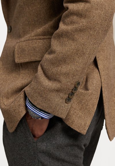 Bruine visgraatjas met een getailleerde pasvorm, voorzien van drie knoopsluitingen, met daaronder een wit en blauw gestreepte shirt en grijze broek zichtbaar.