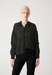 Camisa de renda preta com gola e botões. Apresenta um padrão floral, mangas longas e um corte solto. Combinada com calças pretas.