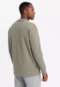 Camisa de manga larga en verde oliva, fabricada en algodón. Presenta un escote redondeado, un corte relajado y detalle de etiqueta en el lateral. Textura suave y lisa.