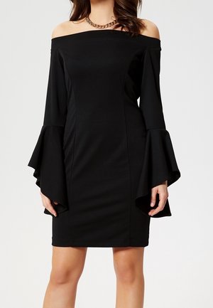 Kvinde iført en sort off-shoulder kjole med flagrende lange ærmer og en chunky guldkæde, stående foran en ensfarvet baggrund.