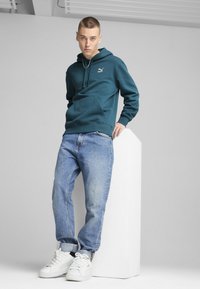 Felpa color teal con tasca frontale e logo bianco, abbinata a jeans azzurri e sneaker bianche. Il modello è in piedi contro uno sfondo grigio chiaro.