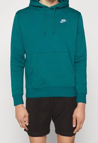 Tyrkysová mikina Nike s kapucí, přední kapsou, šňůrkami na kapuci a malým bílým logem na hrudi; nošena s černými síťovanými šortkami.