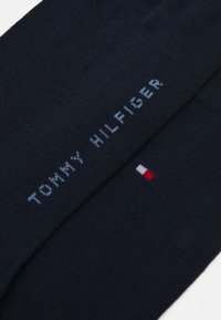 Dunkelblauer Stoff mit "TOMMY HILFIGER" in hellblau und einem kleinen rechteckigen Logo in Rot, Weiß und Navy, gestickt.