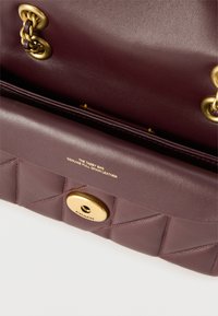 Καφέ δερμάτινη τσάντα χειρός με χρυσά λουράκια αλυσίδας, χρυσό κούμπωμα με στροφή χαραγμένο με τη λέξη "Coach" και εσωτερική ετικέτα που αναγράφει "The Tabby Bag Genuine Full Grain Leather".