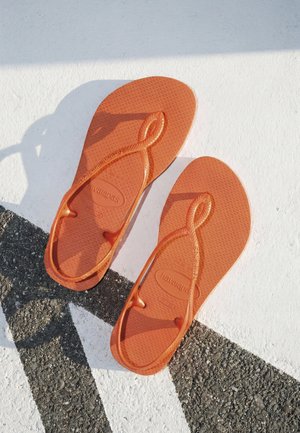 Orange flipflops med teksturerede såler placeret på en hvid overflade med en sort diagonal linje og skygger.