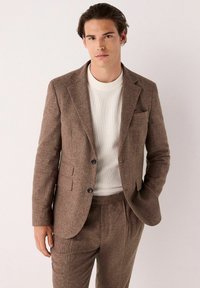 Chaqueta de traje marrón texturizada con dos botones, solapas tipo muesca y un bolsillo en el pecho, combinada con un suéter blanco texturizado claro.